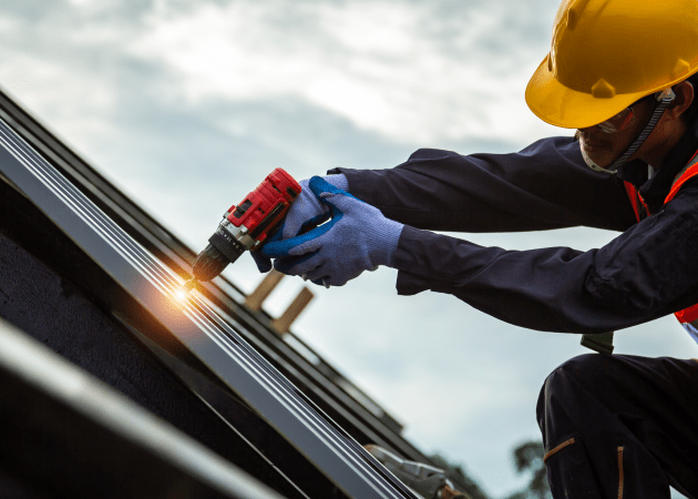 Worker using a power drill on a roof, wearing a yellow hard hat and blue gloves, with sunlight reflecting off the drill.