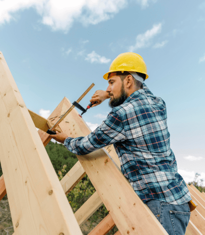 Man wearing a yellow hard hat and plaid shirt using a clamp to secure wooden beams during construction.