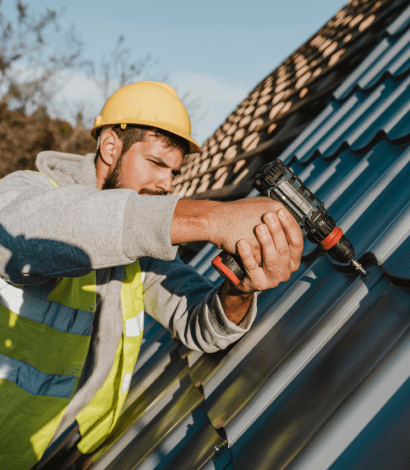 Construction worker in yellow hard hat and reflective vest using a power drill on metal roofing.