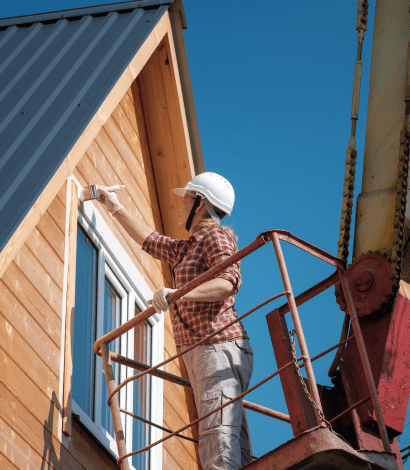 Worker painting a wooden house exterior from a lift, wearing a safety helmet and gloves, with clear blue sky in the background.